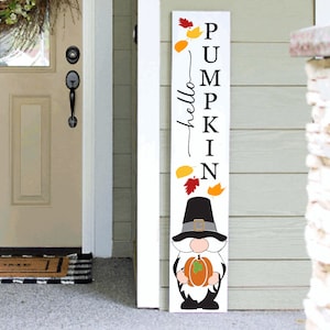 Puede incluir: Un letrero de madera blanco con las palabras "hello pumpkin" y un gnomo sosteniendo una calabaza. El letrero está decorado con hojas de otoño en tonos rojos, naranjas y amarillos.