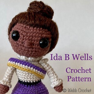 May include: Crochet pattern for a brown-haired doll with a purple dress and a white crocheted top with a gold and purple sash. The doll is named Ida B. Wells. The pattern is by Webb Crochet.