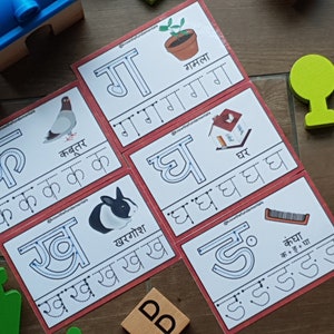 May include: Five red and white flashcards with Hindi alphabet letters and illustrations. The letters are 'क', 'ख', 'ग', 'घ', and 'ङ'. The illustrations are a pigeon, a rabbit, a potted plant, a house, and a comb.