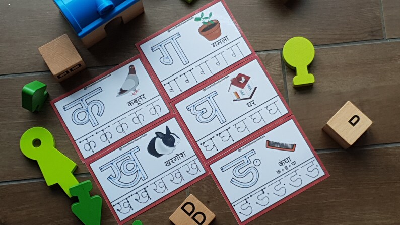 May include: Five red and white flashcards with Hindi alphabet letters and illustrations. The letters are 'क', 'ख', 'ग', 'घ', and 'ङ'. The illustrations are a pigeon, a rabbit, a potted plant, a house, and a comb.
