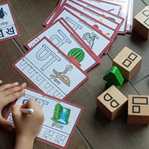 May include: A child is learning the Hindi alphabet with flashcards. The cards feature the letter 'र' (ra) with a picture of a rabbit and the word 'खरगोश' (rabbit) written in Hindi. The child is also using a blue marker to trace the letter on a separate card. There are wooden blocks with letters on them in the background.