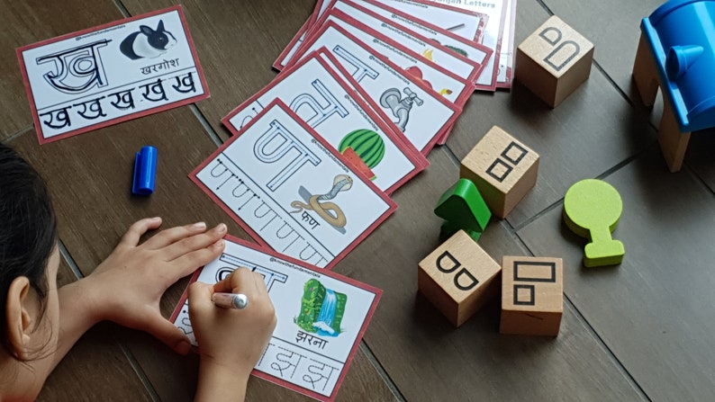 May include: A child is learning the Hindi alphabet with flashcards. The cards feature the letter 'र' (ra) with a picture of a rabbit and the word 'खरगोश' (rabbit) written in Hindi. The child is also using a blue marker to trace the letter on a separate card. There are wooden blocks with letters on them in the background.
