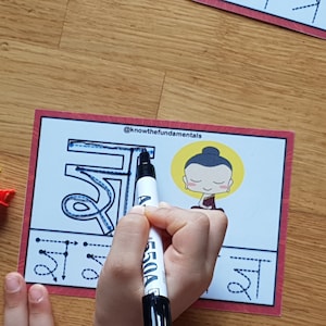 May include: A child traces the Devanagari script for the letter "sha" with a blue marker. The letter is surrounded by dotted lines to guide the tracing. A cartoon illustration of Buddha is in the top right corner of the card. The card is red with white text that reads "@knowthefundamentals".