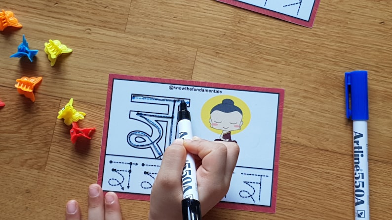 May include: A child traces the Devanagari script for the letter "sha" with a blue marker. The letter is surrounded by dotted lines to guide the tracing. A cartoon illustration of Buddha is in the top right corner of the card. The card is red with white text that reads "@knowthefundamentals".