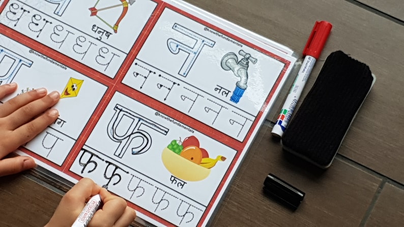 May include: A white dry-erase board with red borders and a child's hand writing the Hindi letter 'फ' (pha) with a red marker. The board has several sections with different Hindi letters and pictures of objects that start with those letters. The words 'फल' (phal) and 'नल' (nal) are written on the board. A black dry-erase marker and a red dry-erase marker are on the table next to the board.