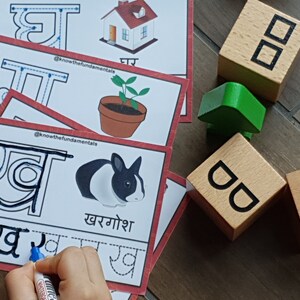 May include: A child's hand traces the Hindi letter 'र' (ra) on a white card with red borders. The card features a black and white rabbit and the word 'खरगोश' (khargoosh) which means rabbit in Hindi. The card also includes a dotted line for tracing the letter.