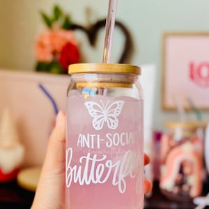 Pu&ograve; includere: Un barattolo di vetro con un coperchio di legno e una cannuccia di vetro trasparente. Il barattolo contiene un liquido rosa e ghiaccio. Il barattolo ha una farfalla bianca e il testo "Anti-Social Butterfly" su di esso.