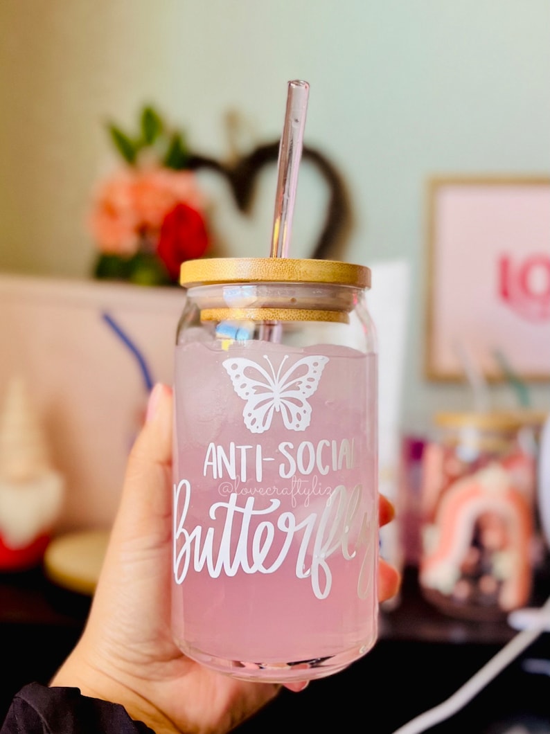 Pu&ograve; includere: Un barattolo di vetro con un coperchio di legno e una cannuccia di vetro trasparente. Il barattolo contiene un liquido rosa e ghiaccio. Il barattolo ha una farfalla bianca e il testo "Anti-Social Butterfly" su di esso.