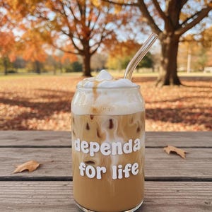 Op de afbeelding: Een glas ijskoffie met een witte schuimtopping en een heldere glazen rietje. Op het glas staat in witte letters "dependa for life". De achtergrond toont herfstbladeren.