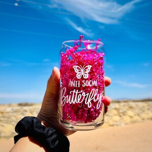 Pu&ograve; includere: Un bicchiere di vetro trasparente con un design a farfalla rosa e il testo "Anti Social Butterfly". Il bicchiere &egrave; pieno di carta crespa rosa.