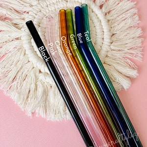 Pu&ograve; includere: Set di sette cannucce da bere in vetro di vari colori, tra cui nero, trasparente, rosa, arancione, verde, blu e turchese. Le cannucce sono disposte a ventaglio su un sottobicchiere di uncinetto bianco.