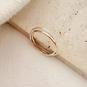Può includere: Un anello composto da due fasce intrecciate, una in tonalità oro lucido e l'altra in tonalità argento. Le fasce presentano un profilo liscio e arrotondato, unite insieme per formare un unico gioiello con una finitura metallica.
