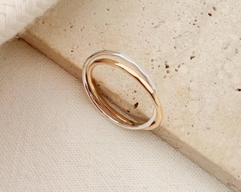 May include: A ring featuring two intertwined bands, one in a polished gold tone and the other in a silver tone. The bands have a smooth, rounded profile and are joined together to form a single continuous piece of jewelry with a metallic finish.