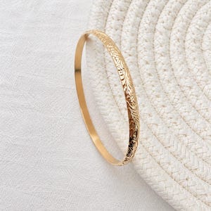 Pode incluir: Uma pulseira rígida circular e fina em ouro, apresentando um intrincado padrão floral em relevo na sua superfície exterior. O metal tem um acabamento polido e reflexivo que capta a luz, realçando as gravações detalhadas na banda.