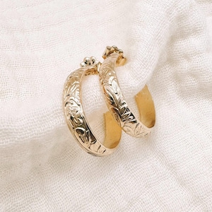 May include: A pair of gold hoop earrings featuring an intricate floral engraving across the outer surface. The earrings have a polished finish and a curved, semi-circular design, showcasing detailed botanical patterns etched into the metal.