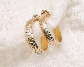 May include: A pair of gold hoop earrings featuring an intricate floral engraving across the outer surface. The earrings have a polished finish and a curved, semi-circular design, showcasing detailed botanical patterns etched into the metal.