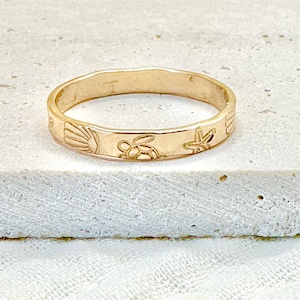 May include: A gold ring with a beach theme. The ring has engraved designs of a seashell, turtle, and starfish. The ring is set against a neutral background.