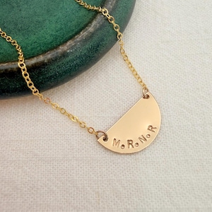 May include: A gold-colored necklace featuring a semi-circular pendant attached to a delicate chain. The flat, curved metal charm is stamped with the letters M, R, N, and R, each separated by a small dot, following the arc of the pendant's edge.