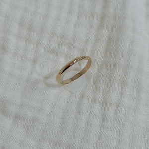 May include: A gold ring with a smooth, rounded band. The ring is set against a white, textured fabric background. The ring appears to be a simple band, possibly a wedding band or stacking ring.