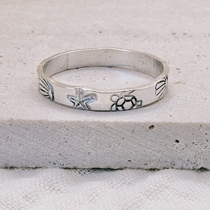 May include: A silver ring with a design of seashells, a starfish, and a turtle. The ring is on a white surface.