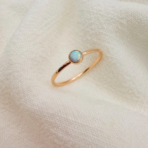 May include: A thin gold-toned ring featuring a single, small, round, iridescent opal gemstone set in a bezel setting. The band is smooth and circular, showcasing a polished metallic finish that reflects light across its surface.