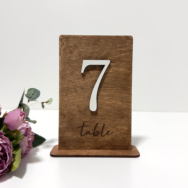 May include: A wooden table number with the number 7 and the word "table" engraved on it. The number is white and the wood is a dark brown color.