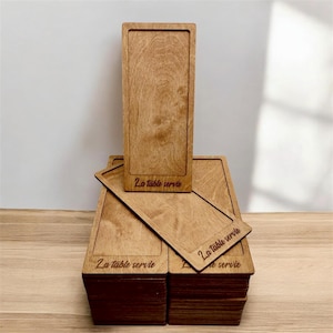 May include: A stack of rectangular wooden serving trays with a light brown finish. Each tray has a recessed area and the phrase "La table servie" is etched on the surface. One tray is standing upright.