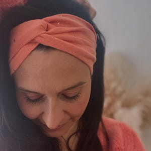 Könnte beinhalten: Ein korallenfarbenes Stirnband mit Goldflocken wird auf dem Kopf getragen. Der Stoff ist oben verdreht. Die Person, die das Stirnband trägt, blickt nach unten. Die Person trägt einen korallenfarbenen Pullover.