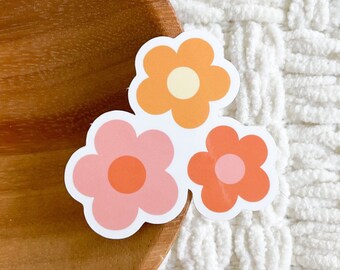 Peut inclure: Trois autocollants de fleurs de style rétro en rose, orange et jaune. Les autocollants sont disposés en grappe sur une surface blanche et duveteuse.