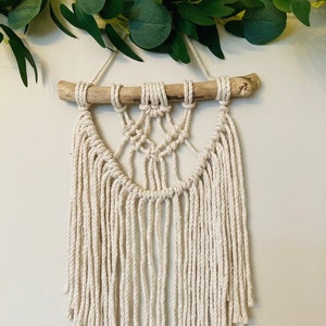 May include: A macrame wall hanging with a natural wood branch and white cotton cord. The hanging features a geometric pattern with fringe.