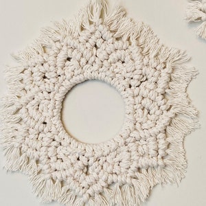 May include: A white macrame wall hanging with a circular opening in the centre and a fringe border.