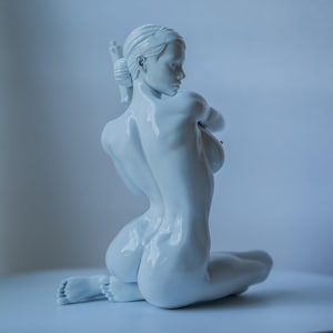 May include: A white sculpture of a kneeling figure. The figure is facing away from the viewer, with their arms crossed in front of them. The sculpture is on a white surface. The figure's hair is pulled back.