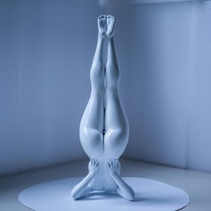 Może przedstawiać: Biała ceramiczna rzeźba osoby w pozie jogi, stojącej na rękach z nogami w powietrzu.
