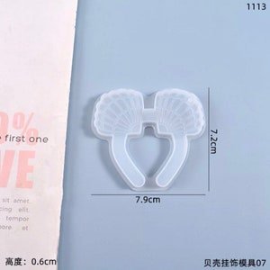 May include: A clear, seashell-shaped mold for crafting. The mold is 7.9 cm wide and 7.2 cm tall, with a thickness of 0.6 cm. The design features two fan-shaped elements connected by a curved base. The text "1113" is visible.