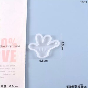 May include: A clear, hand-shaped silicone mold, 6.8 cm wide and 5.5 cm tall, with a small loop for hanging. The mold is on a light blue background. Text in the image reads "the first one" and "VE".