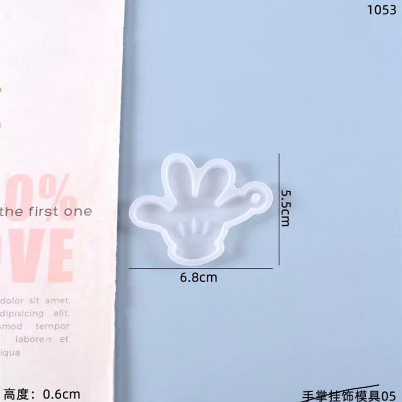 May include: A clear, hand-shaped silicone mold, 6.8 cm wide and 5.5 cm tall, with a small loop for hanging. The mold is on a light blue background. Text in the image reads "the first one" and "VE".