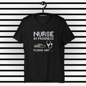 Pode incluir: Camiseta preta com texto branco que diz "Enfermeira em progresso" com uma barra de progresso que diz "Carregando" e um estetoscópio pixelado. O texto abaixo da barra de progresso diz "Por favor, aguarde..."