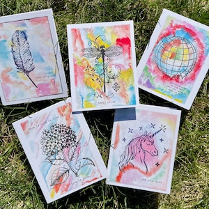 May include: Five watercolor greeting cards with various designs. One card features a feather with the text "loud and proud." Another has a dragonfly with "happy pride." A third shows a globe with text. The others have a flower and a unicorn with text.