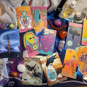 May include: A tarot card spread with 10 cards, including The Chariot, The Magician, Strength, The Emperor, Queen of Chiles, and Deer. The cards are laid out on a purple and black patterned surface with crystals and other objects.
