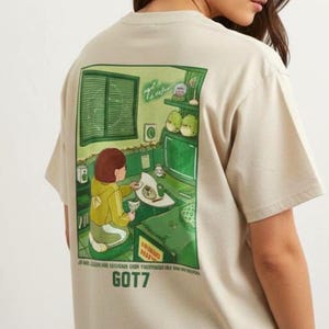 Puede incluir: Camiseta beige con una ilustración verde y blanca en la espalda. El gráfico representa a una persona en una habitación con un ordenador y otros objetos. El texto "GOT7" está impreso debajo de la ilustración. La camiseta tiene mangas cortas.