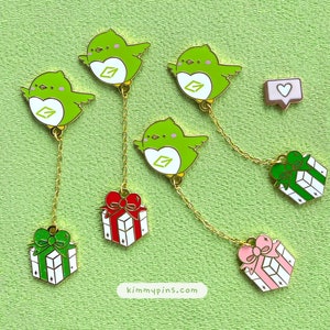 Può includere: Cinque spille smaltate con un uccello verde con un logo a forma di cuore e una scatola regalo appesa a una catena dorata. Le scatole regalo sono rosse, verdi e rosa. Le spille sono su uno sfondo verde. Il testo "kimmypins.com" è visibile nella parte inferiore centrale dell'immagine.