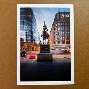 May include: A city street scene featuring a bronze equestrian statue with a red hat. Modern buildings are on either side of the statue, and a blurred bus passes by. The sky is a mix of blue and orange.