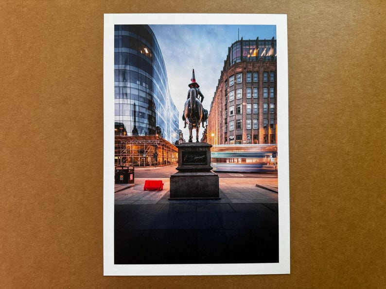 May include: A city street scene featuring a bronze equestrian statue with a red hat. Modern buildings are on either side of the statue, and a blurred bus passes by. The sky is a mix of blue and orange.