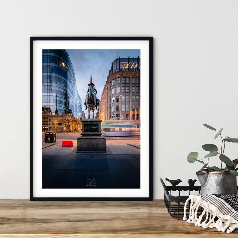 May include: Framed photograph of a city scene. A bronze equestrian statue is in the foreground, with modern glass buildings and a traditional brick building behind. A red traffic barrier is visible.