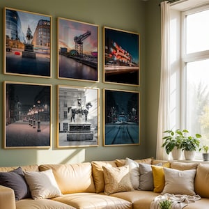 May include: Six framed art prints with gold frames hang on a green wall above a tan sofa. The prints depict cityscapes, including a statue, a bridge, and a building with bright lights. The room has a large window with plants.