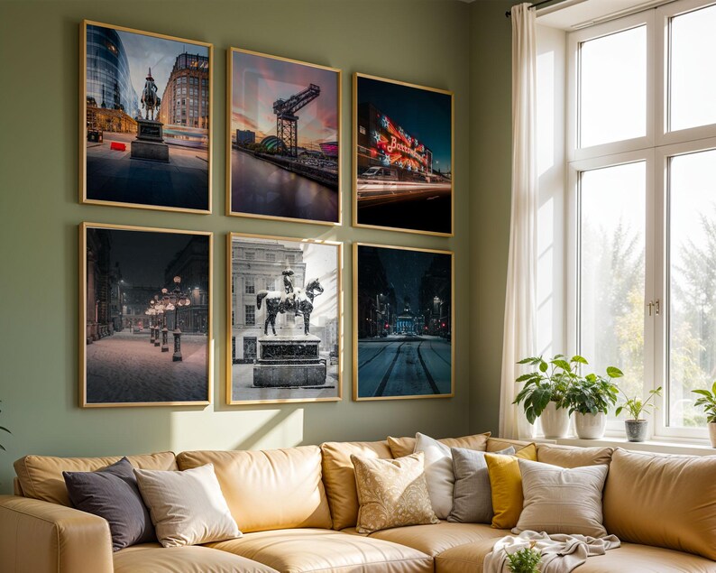 May include: Six framed art prints with gold frames hang on a green wall above a tan sofa. The prints depict cityscapes, including a statue, a bridge, and a building with bright lights. The room has a large window with plants.
