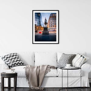 May include: A framed photograph of a city scene at dusk. The image features a statue of a person on a horse in front of buildings. The sky is a mix of blue and orange, and the buildings have lights on.