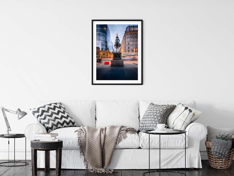 May include: A framed photograph of a city scene at dusk. The image features a statue of a person on a horse in front of buildings. The sky is a mix of blue and orange, and the buildings have lights on.