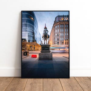 May include: Framed photograph of a city street scene featuring a bronze equestrian statue with a red traffic cone on the rider's head. Modern buildings and a bus are in the background, with a red object on the pavement.