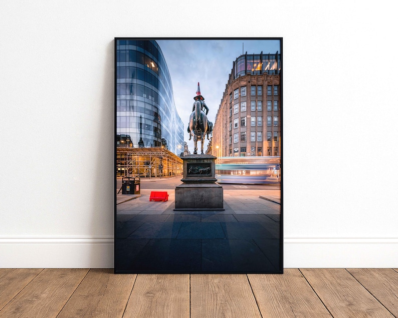 May include: Framed photograph of a city street scene featuring a bronze equestrian statue with a red traffic cone on the rider's head. Modern buildings and a bus are in the background, with a red object on the pavement.